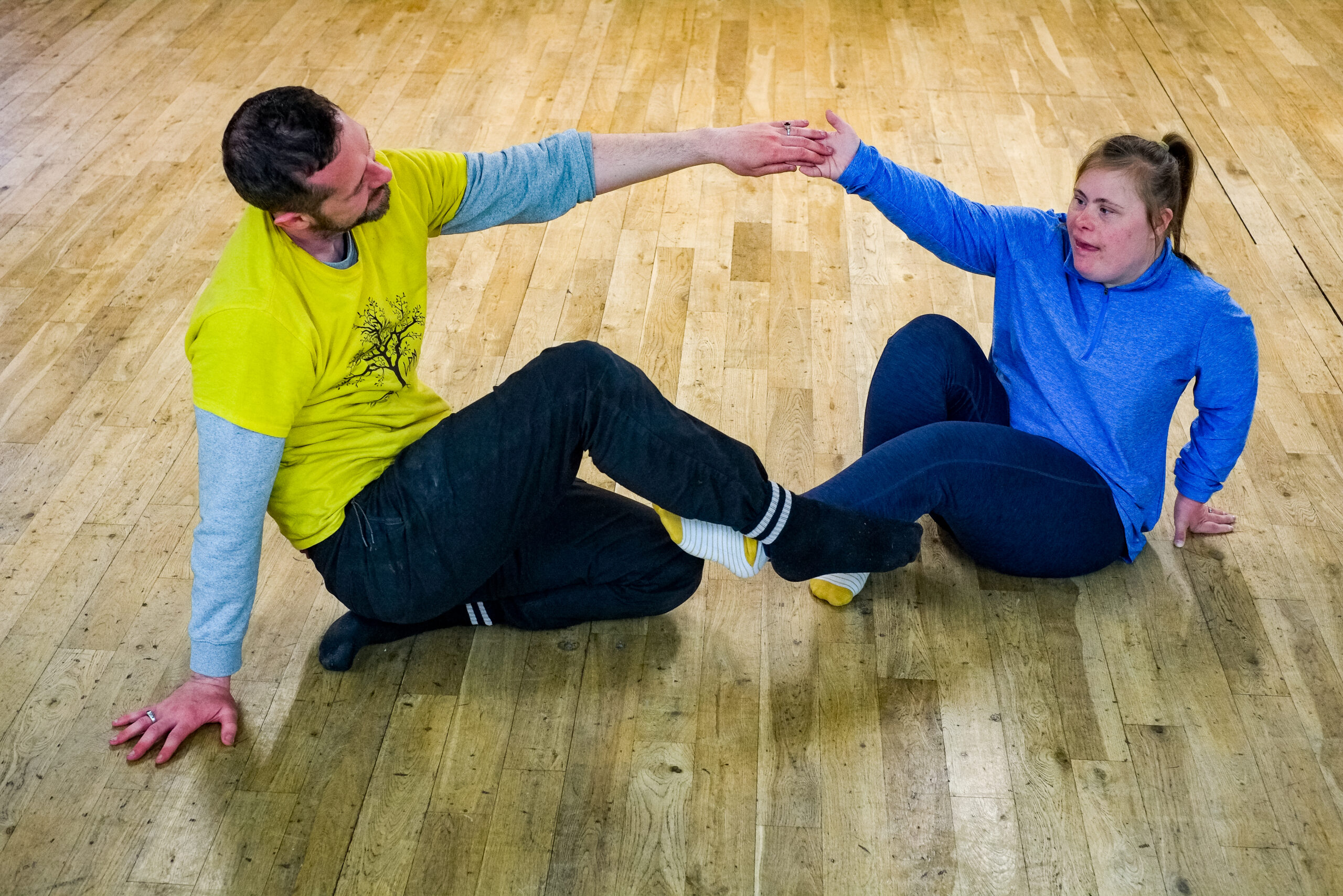 George, a white male dancer wearing a yellow t-shirt, sits opposite Leanne, a white female participant dancer with Downs Syndrome wearing blue. George and Leanne hold hands.
